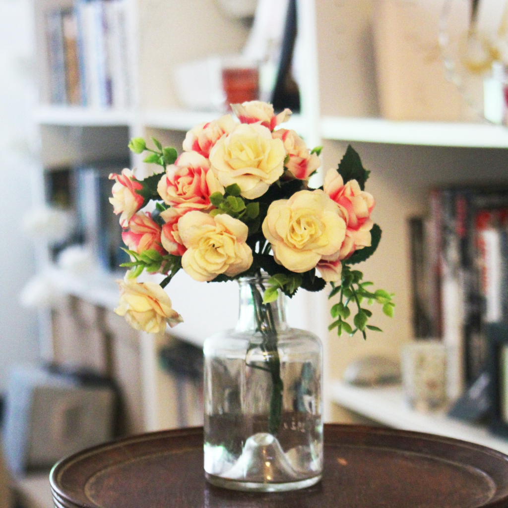 Conjunto de flores artificiales sobre un jarrón en una mesita de una biblioteca o una habitación.