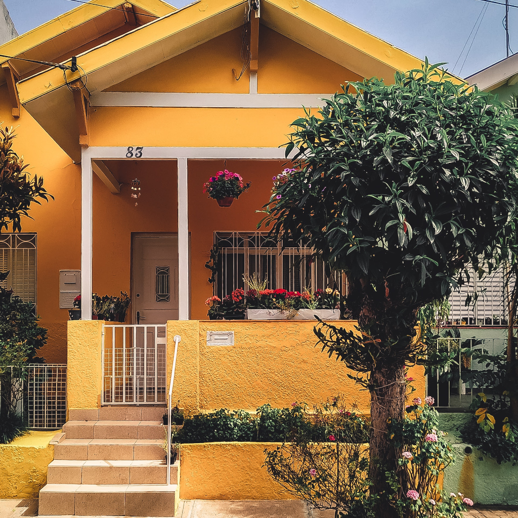 Fachada de una casa pintada de color amarillo con muchas plantas artificiales y un árbol no natural.